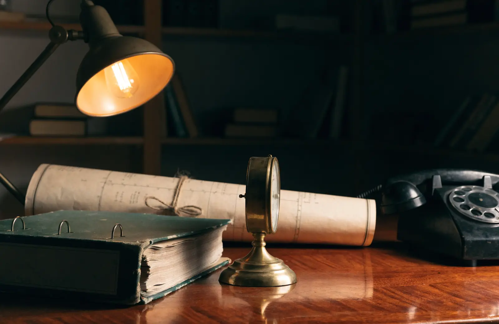 A ring binder, brass ship's clock and rolled chart on a mahogany desk under a lamp